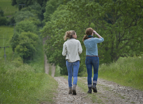Two young women walking down a path, fields to the side of them with hedges and trees lining them. One woman is looking through binoculars