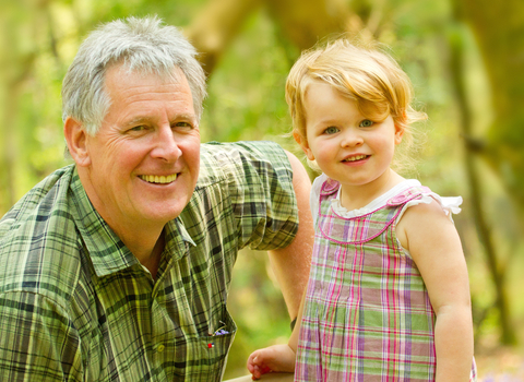 A grandfather and his toddler granddaughter in a woodland, smiling towards the camera