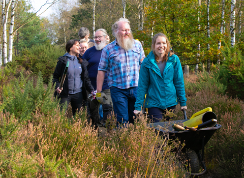 A group of volunteers walking through heath at a nature reserve, pushing a wheelchair and carrying tools
