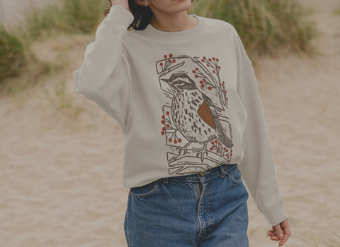 A woman on a beach wearing a beige sweatshirt with a redwing graphic design