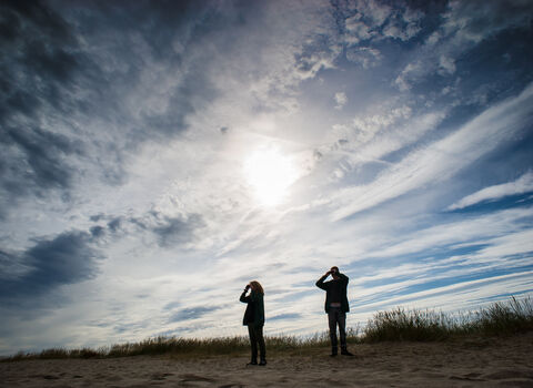 Two people stood on a beach with sun and clouds above them, looking out through binoculars