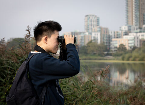 A young man standing with binoculars, looking over a wetland for birds. In the background high rise buildings can be seen