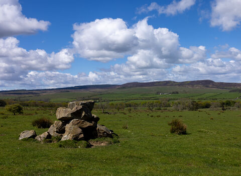 Wide landscape view of the Simonside Hills on the Rothbury Estate, with a pile of weathered rocks in the foreground and rolling green moorland stretching into the distance. The hills rise along the horizon beneath a bright blue sky filled with large white clouds.