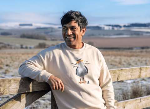 A man leaning on a fence wearing a jumper featuring a gull wearing sun glasses and eating chips
